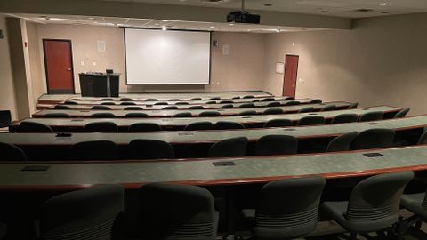 Lecture room with several raised rows of tables and chairs facing a projector screen and podium
