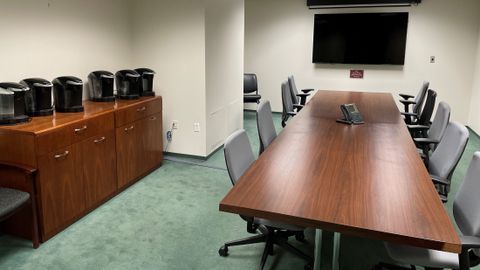 A long table with multiple chairs, desk phone, TV monitor and Keurigs sit in a conference room